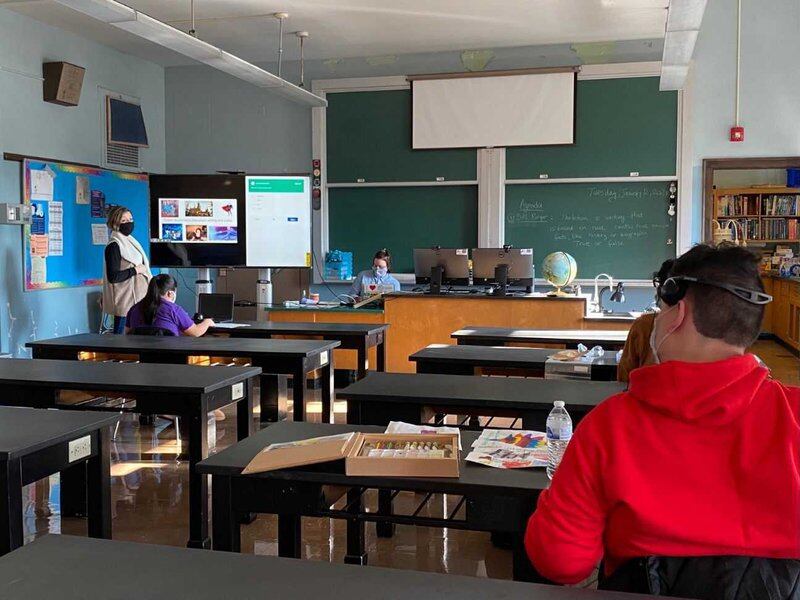 A teacher and students several feet apart sit at desks, working in a classroom