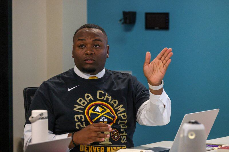 Denver school board Vice President Aoun’tai Anderson gestures while speaking at a table during a school board meeting. He’s a young Black man wearing a Nuggets 2023 NBA Championship t-shirt over a white dress shirt.