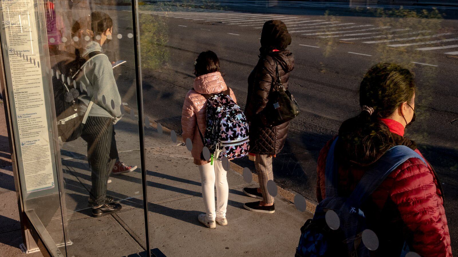 A woman stands with her two daughters and another woman at a city bus stop.
