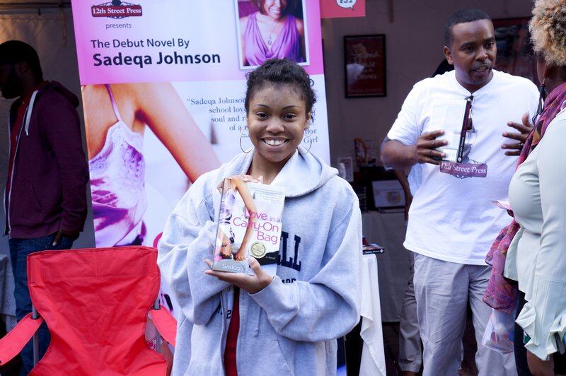 A woman wearing a hooded sweater smiles and holds up a book in front of a banner saying “The Debut Novel by Sadeqa Johnson.”