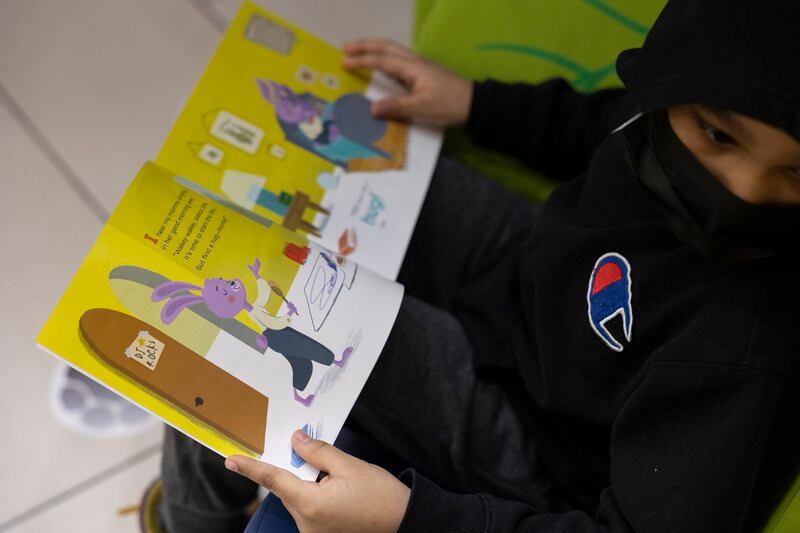A young boy in a black Champion hoodie and mask reads a children’s book.
