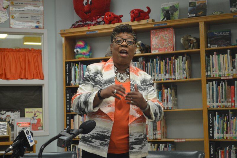 A woman wearing an orange and black jacket stands behind a microphone set up in a library.