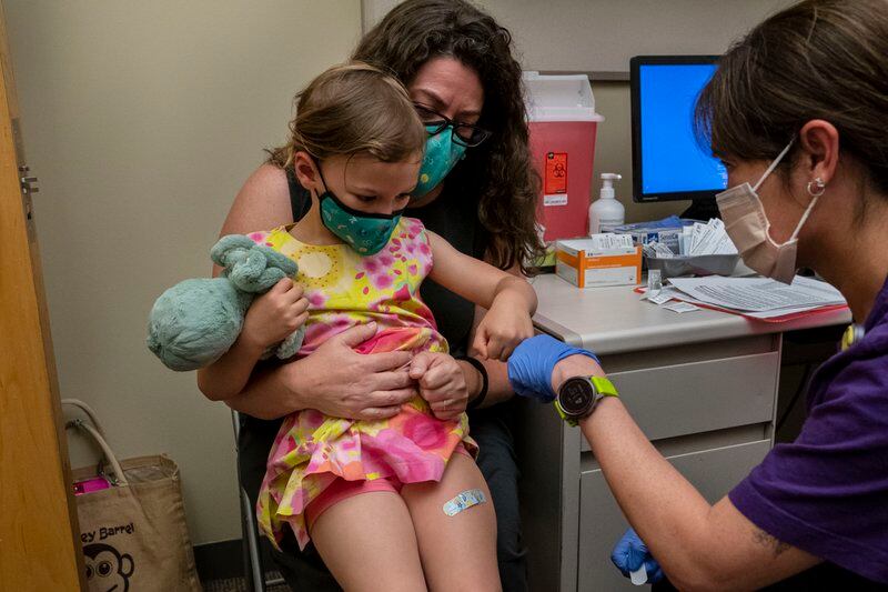 A young child being held by her mother receives a vaccine from a health care professional.