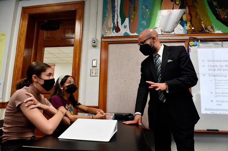 Two masked students sit at desks. Superintendent Alex Marrerro, wearing a mask and a suit, stands and talks with them.