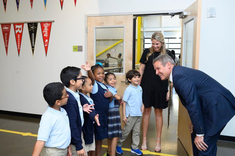 Man in a suit bends down to talk to six elementary students lined up outside a room.