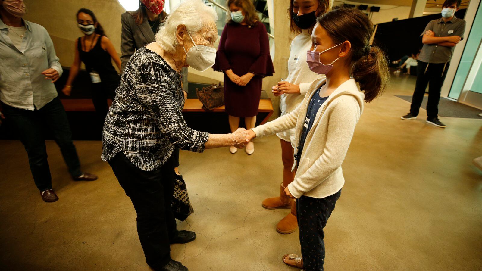 An elderly woman shakes hands with a girl and they make eye contact both wearing masks