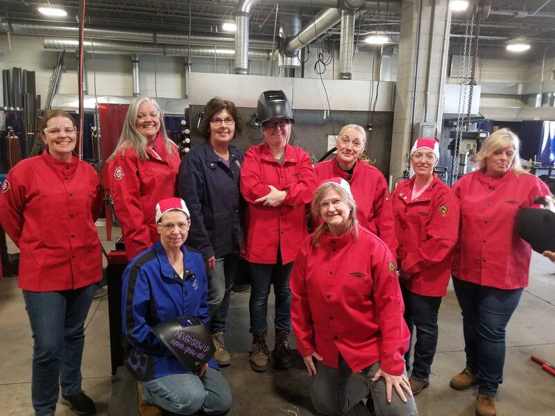 Nine women, mostly in red welding jackets, pose for a photo in a welding shop.