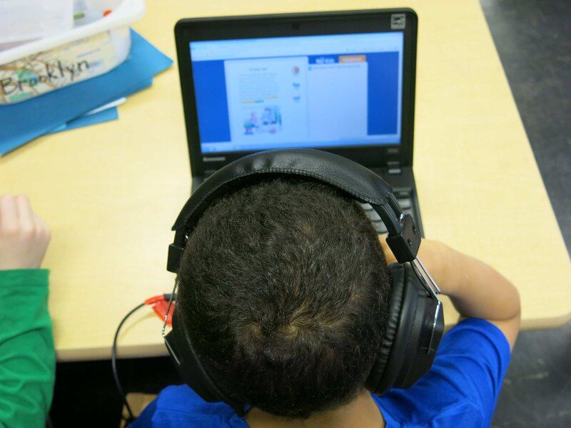 A young student wearing headphones and working on a laptop. The foreground is a close up of the child’s head with the laptop in the background.