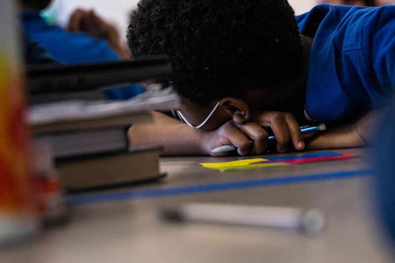 A student puts their head on a desk with a pencil in his hand. A pile of books is seen off to one side.