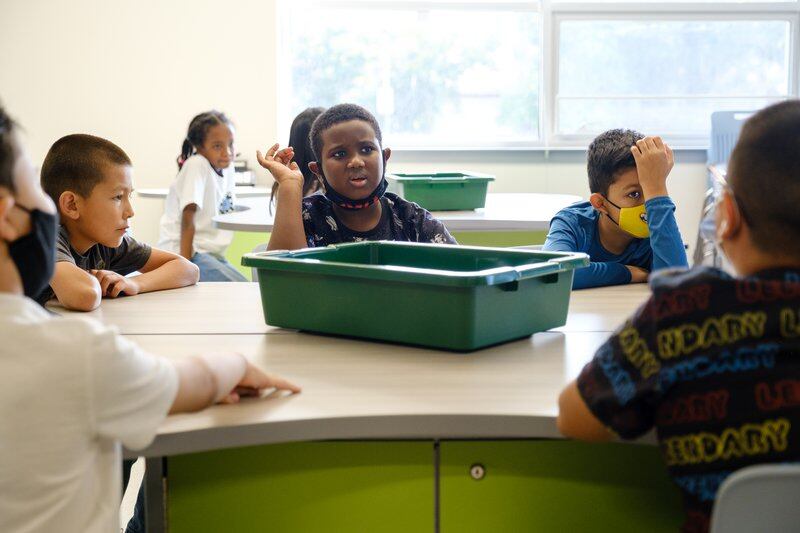 Elementary students with and without masks sit around a table with a green plastic bin in the middle of the table.