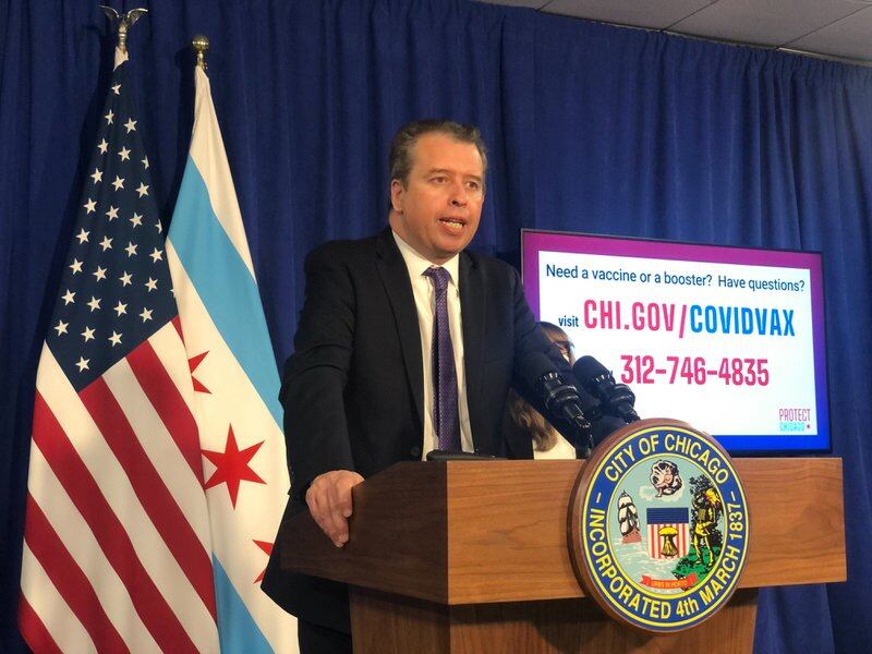 Chicago Public Schools CEO Pedro Martinez, wearing a black suit and purple tie, speaks at a wooden City of Chicago podium during a press conference. There are Chicago and United States flags hanging behind him against a blue backdrop, along with a screen providing COVID information.