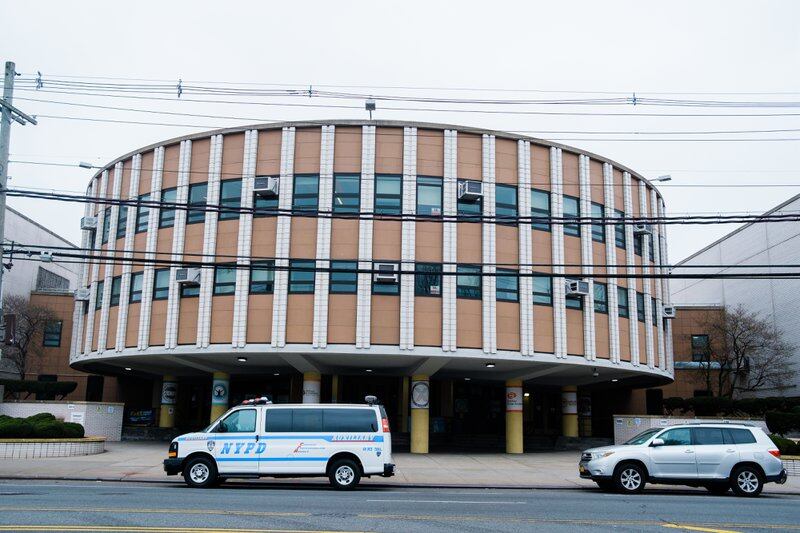 The curved facade of Brooklyn High School for Excellence and Equity in Canarsie. There is an NYPD van parked outside, as well as a standard silver SUV.