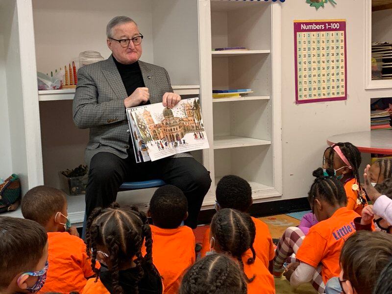 A man wearing glasses and black pants holds a book out for children sitting on the floor and wearing orange shirts to see.
