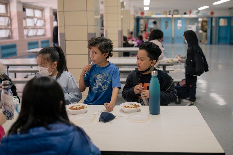Three young students at P.S. 020 Anna Silver in New York City eat school lunch with others at a cafeteria table.