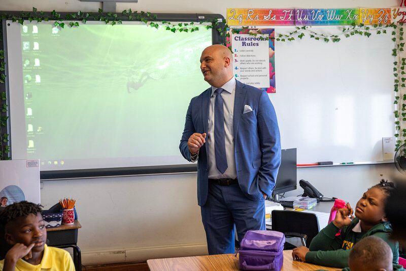 Man wearing a blue suit and tie at the front of a school classroom.