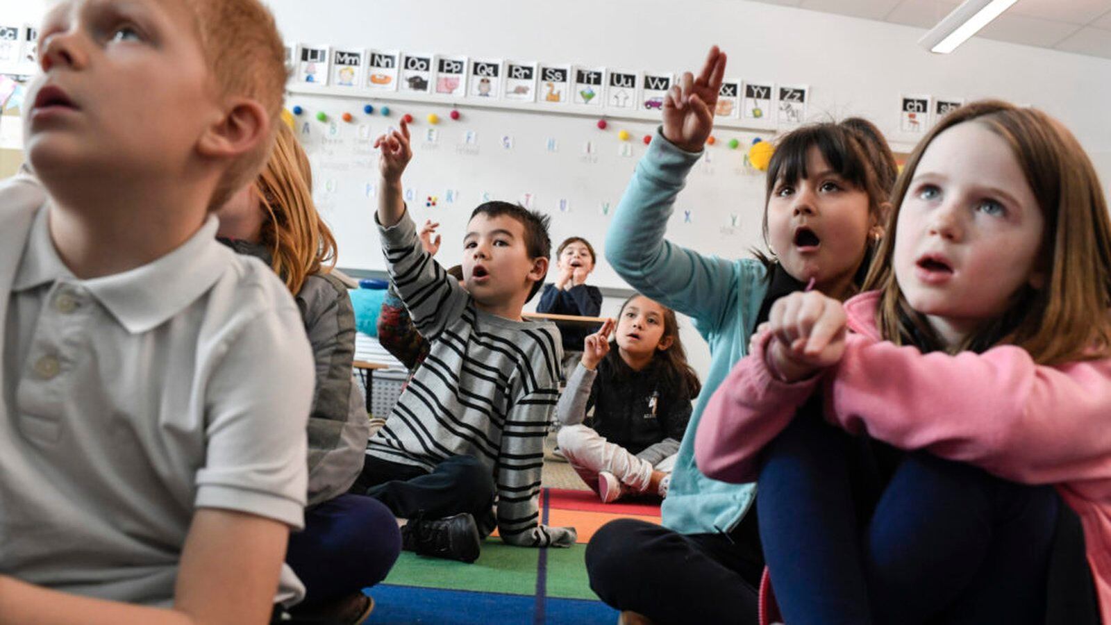 First-graders sit on a rug in a classroom. Their hands are in the air, tracing letters. Their mouths are open as they repeat after the teacher.