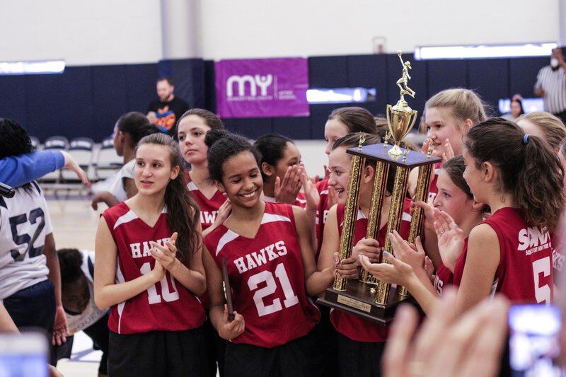 A group of girls wearing red uniforms hold a large old trophy.