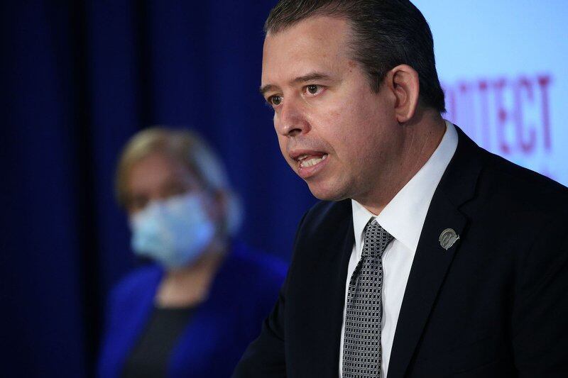 Chicago Public Schools CEO Pedro Martinez speaks during a press conference. A woman wearing a blue pantsuit and surgical mask looks on in the background.