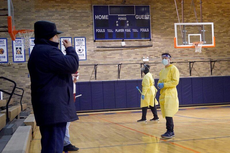 A man uses a nasal swab COVID test in a high school gymnasium, as two people in yellow protective suits and masks stand before him on the hardwood floor.