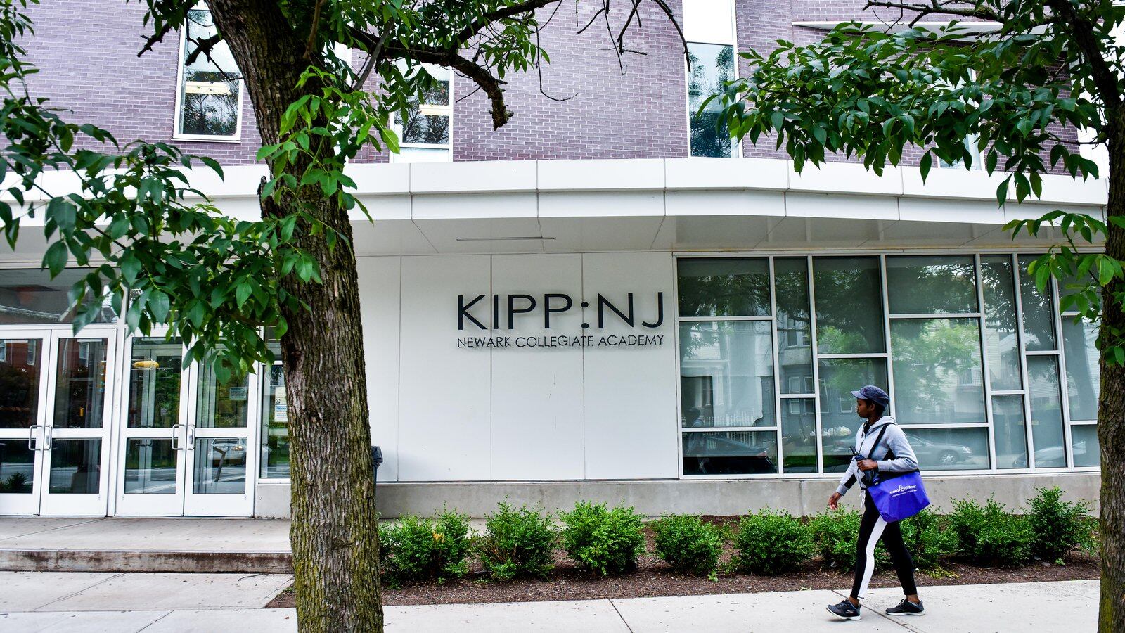A student wearing a cap and carrying a blue tote bag walks on a walkway in front of the KIPP-NJ building.