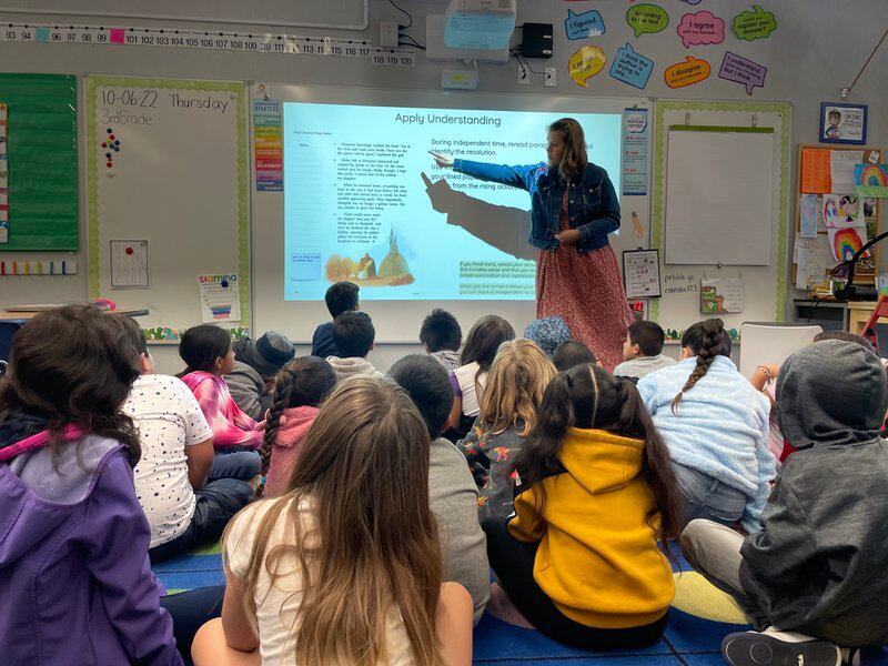 A teacher points to information on a white board as her students sit on the floor in front of her.
