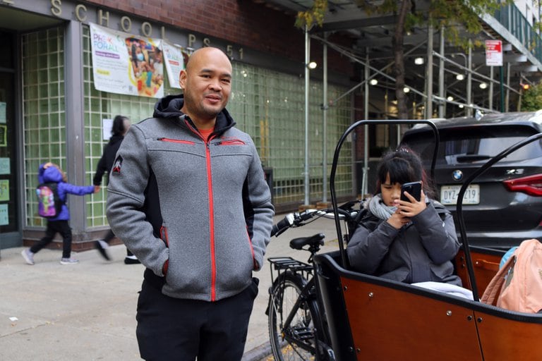 A man with a grey jacket smiles at the camera next to his young daughter who is holding a cellphone with a green brick building in the background.