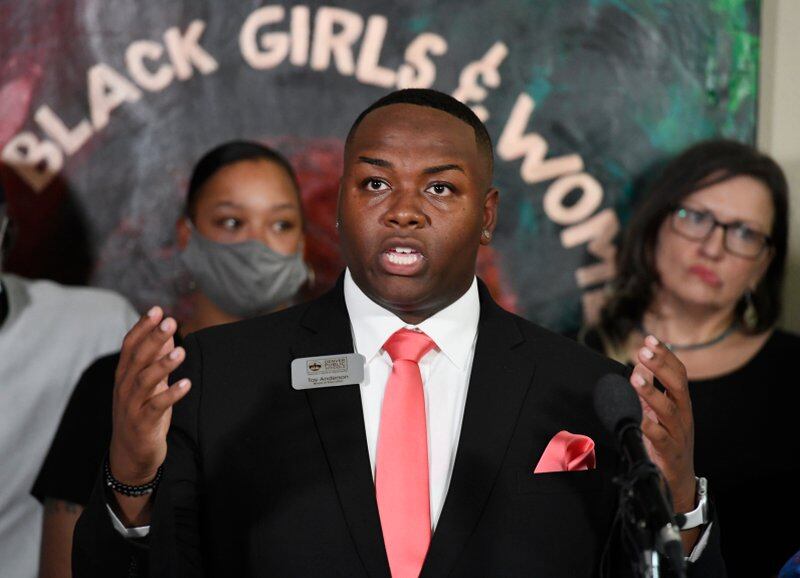 Denver school board member Tay Anderson gestures while speaking at a press conference. He is wearing a black suit and salmon colored tie. Supporters are standing behind him.