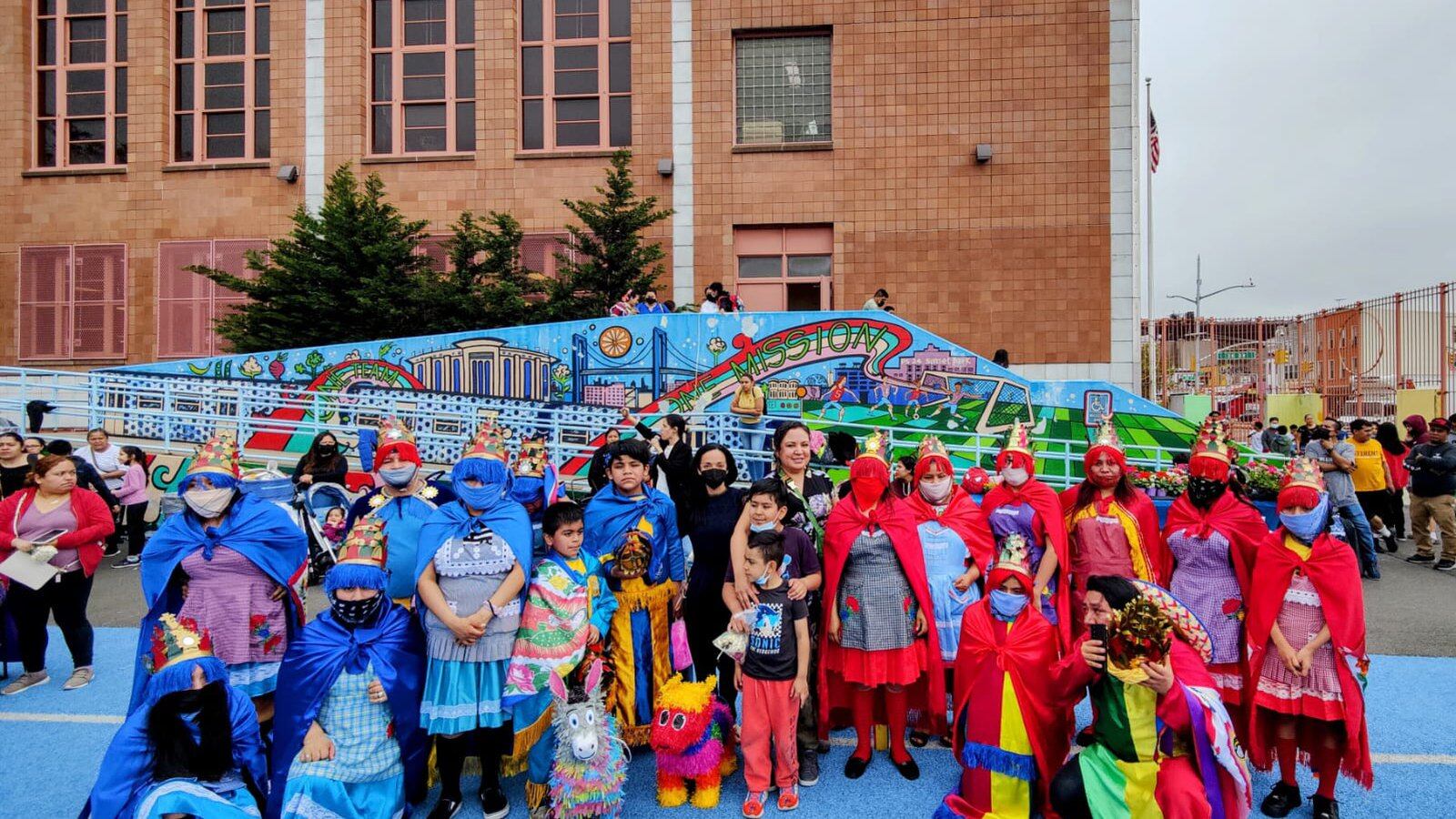 Parents and students in bright blue and red costumes line up in front of a school mural outside of a brick building.