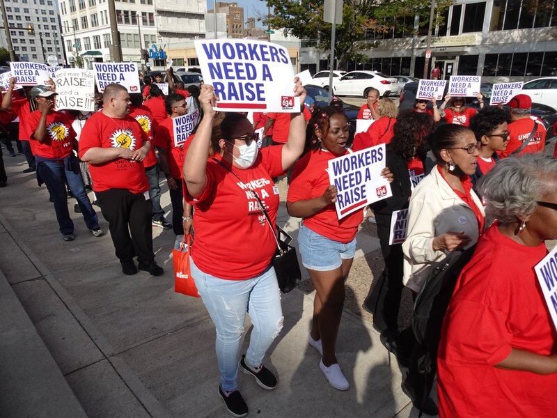 A group of people in red shirts hold signs that say “workers need a raise.”