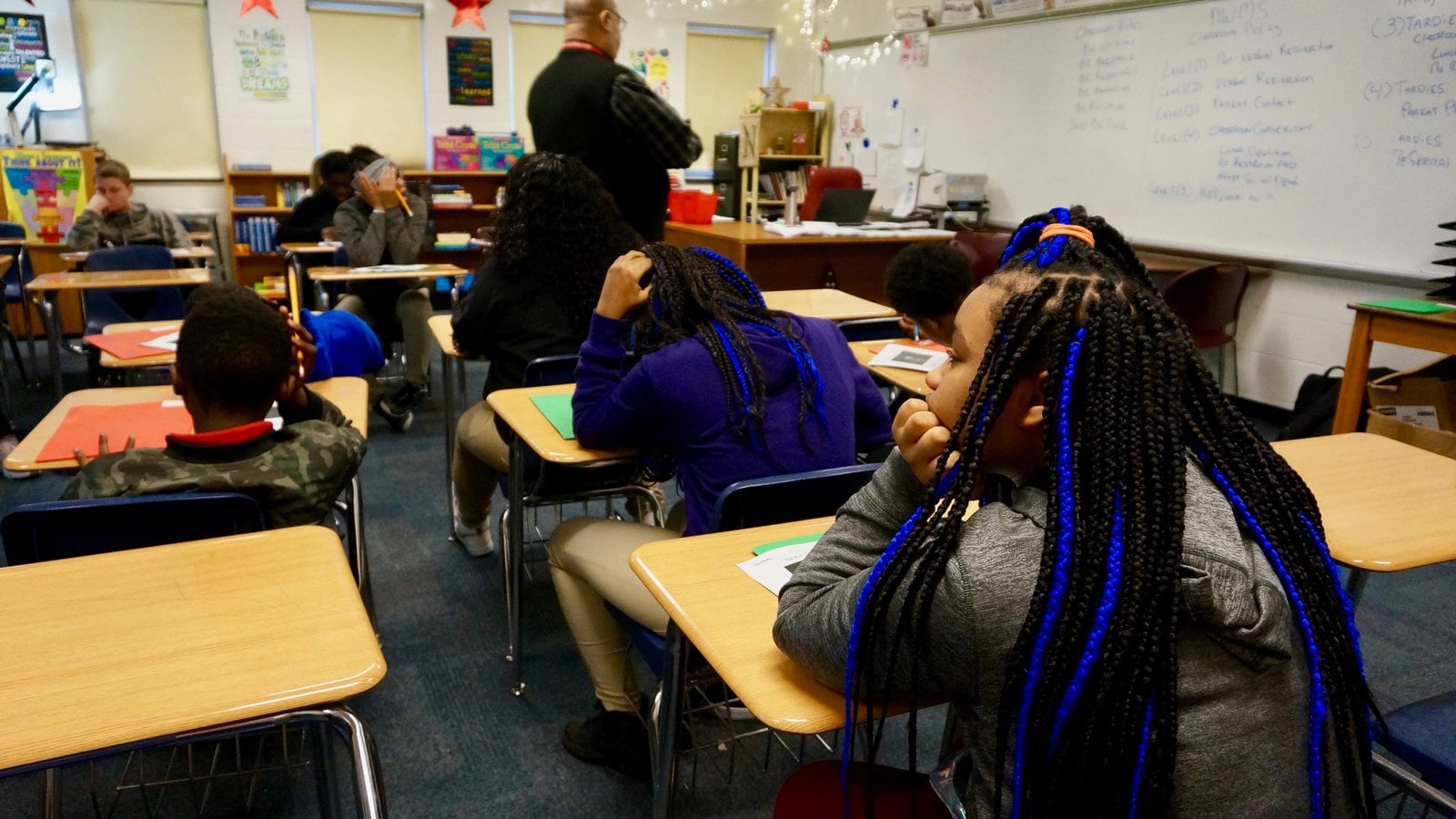 Three students in the foreground sit at desks in a classroom with their backs to the camera. A teacher walks through a line of desks in the background.