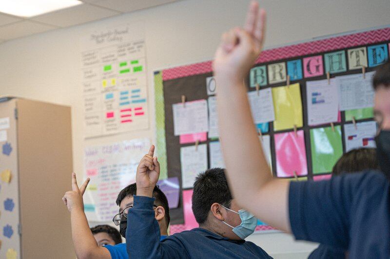 Students raise their fingers in a classroom together, all wearing protective masks.