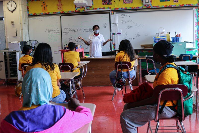 A person in a white shirt stands in front of a white board in a classroom. Students sit in desks in front of the person.
