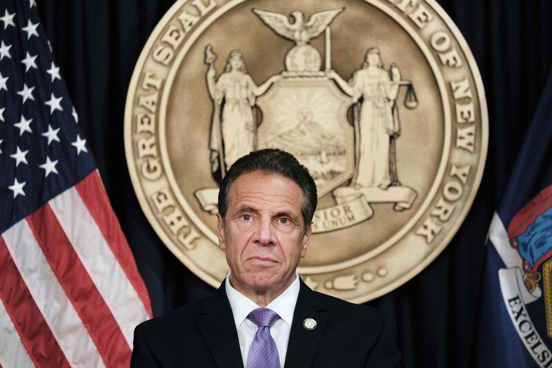 New York Governor Andrew Cuomo sits in front of the seal of the state of New York behind him, between the flags of the United States and New York.