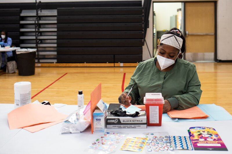 woman in white mask and headband with green shirt sits at table with syringes and medical supplies in front of her