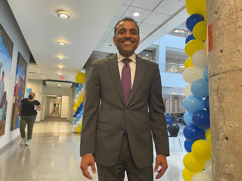 A smiling man in a gray suit stands in a hallway as a woman walks behind him. There are blue, yellow, and white balloons to his left.