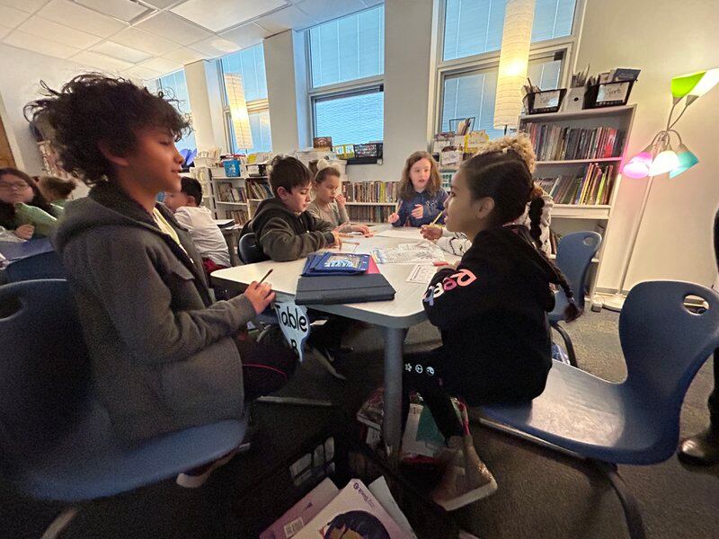 Six elementary students sit talking at a table with papers and pencils.