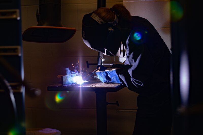 A young woman welds a joint, sparks flying from her workstation.
