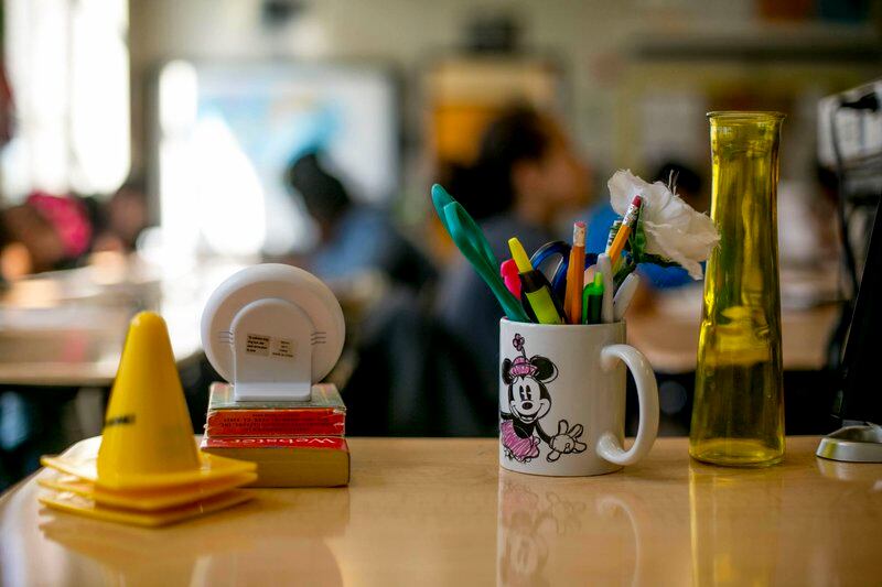 Markers, pencils and pens in a Minnie Mouse coffee cup, small yellow cones, a dictionary, and other items sit atop a desk in a classroom.