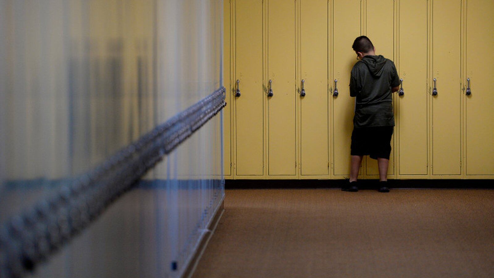 A wide hallway ends in a bank of yellow lockers. A boy who looks about 11 or 12, with short hair and a black T-shirt and shorts, is seen from behind. He’s opening one of the lockers. He’s alone.