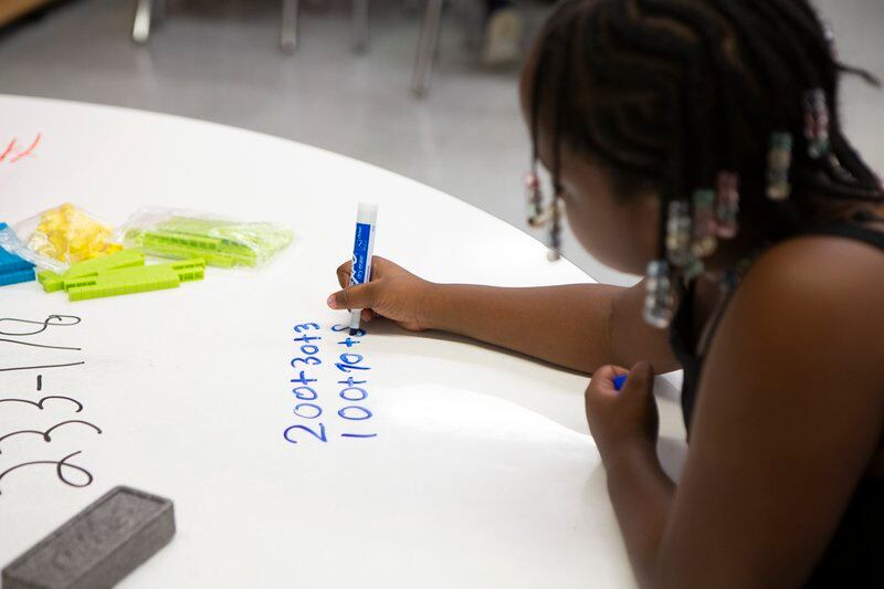 A young girl holds a blue marker in her hand as she sits at a desk and writes an equation on poster paper.