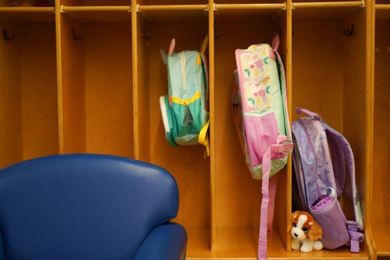 A pale blue backpack and a pink backpack hang in adjacent vertical wooden cubbies; next to them a lavender backpack sits in another cubby with a small stuffed dog. Part of a royal blue cushioned chair shows in one corner of the foreground.