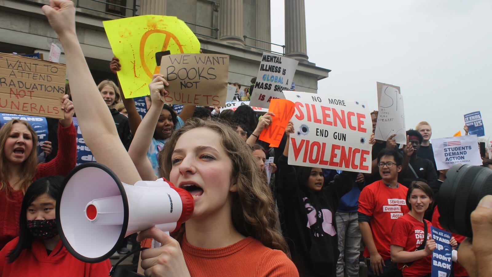 A girl yells into a megaphone surrounded by students holding signs on the steps of a large building.