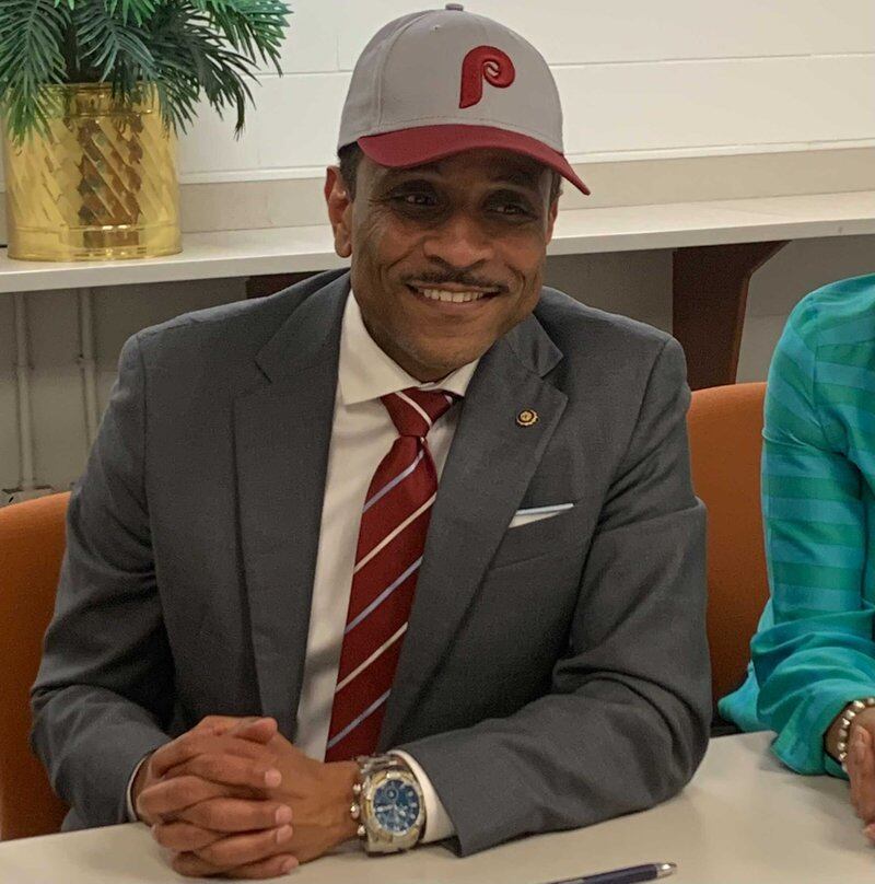 A man in a red and white tie and gray suit is wearing a Philadelphia Phillies hat while seated at a table.
