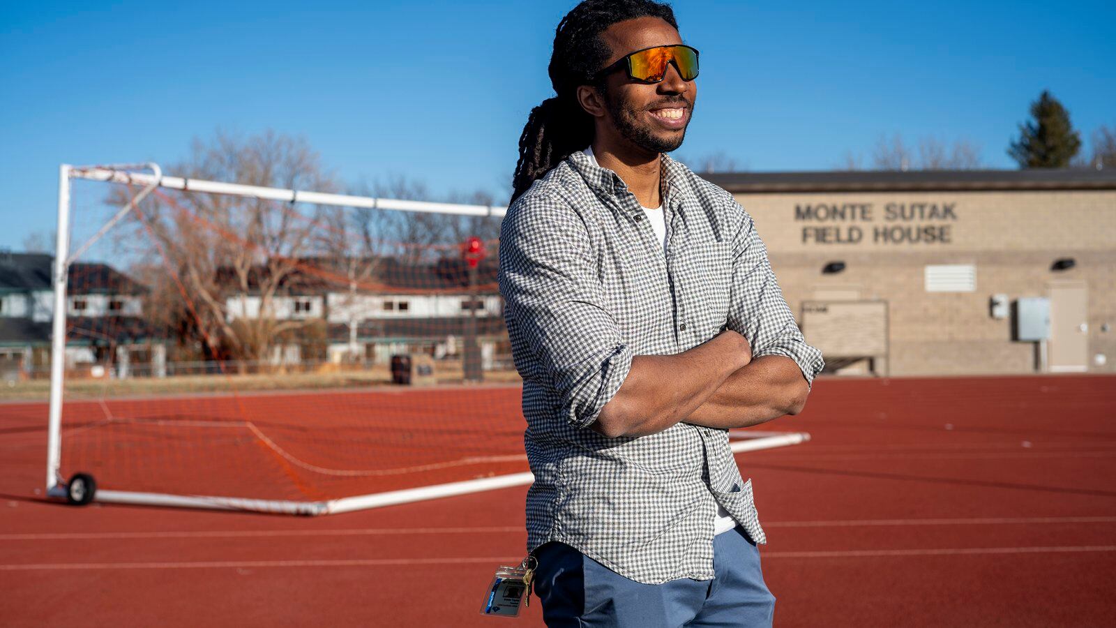 A young, Black educator and track coach, wearing a button-up shirt, and bright multi-colored sunglasses, smiles as he stands on the edge of the track.