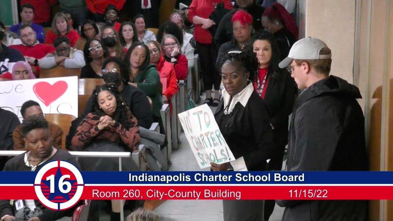 people in chairs at a meeting and one woman holds a sign that says no to charter schools.