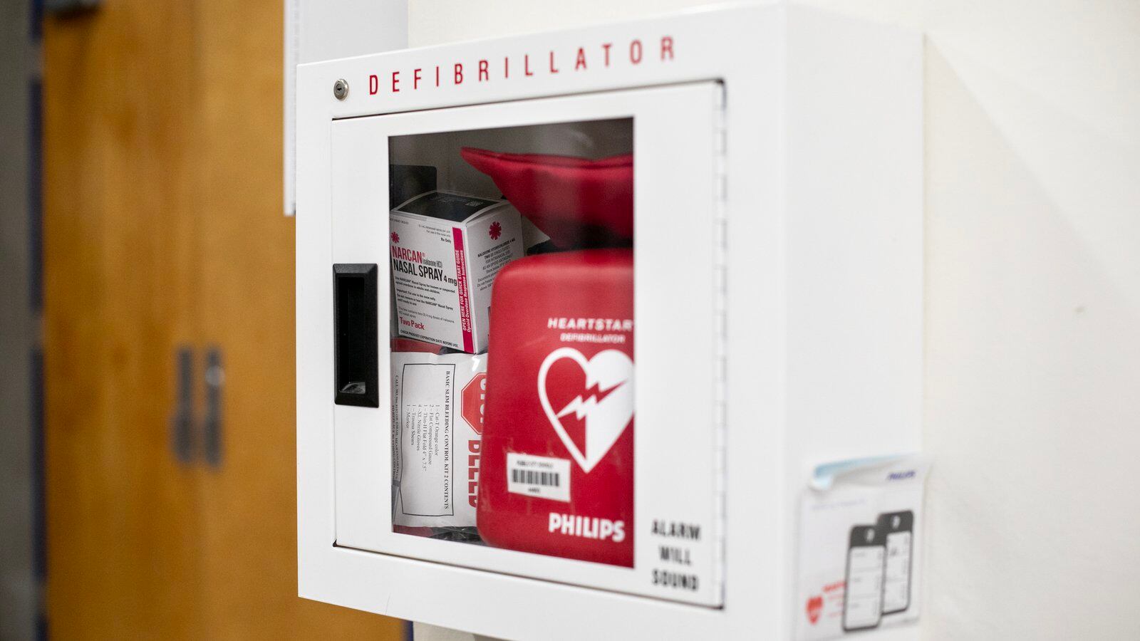 An emergency box mounted in the hallway of a building, containing a defibrillator and medications.