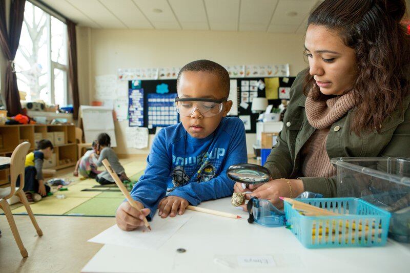 A teacher sits at a table with a kindergarten boy who is looking at a paper using a magnifying glass.