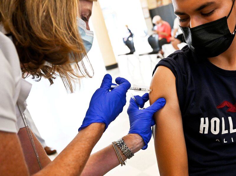A student wearing a Hollister shirt receives a dose of a COVID vaccine from a health care professional wearing purple latex gloves.