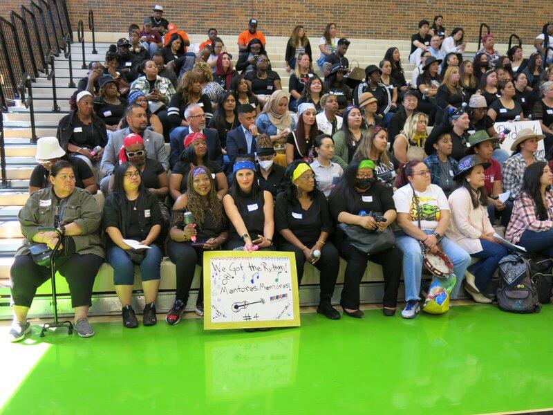 A group of people sit in the bleachers at Harry S Truman Community College in Chicago. Holding signs along a bright green floor.