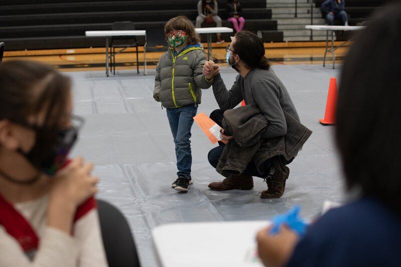 A young boy wearing a winter coat is comforted by an adult kneeling next to him before getting a vaccine shot inside a school gym.